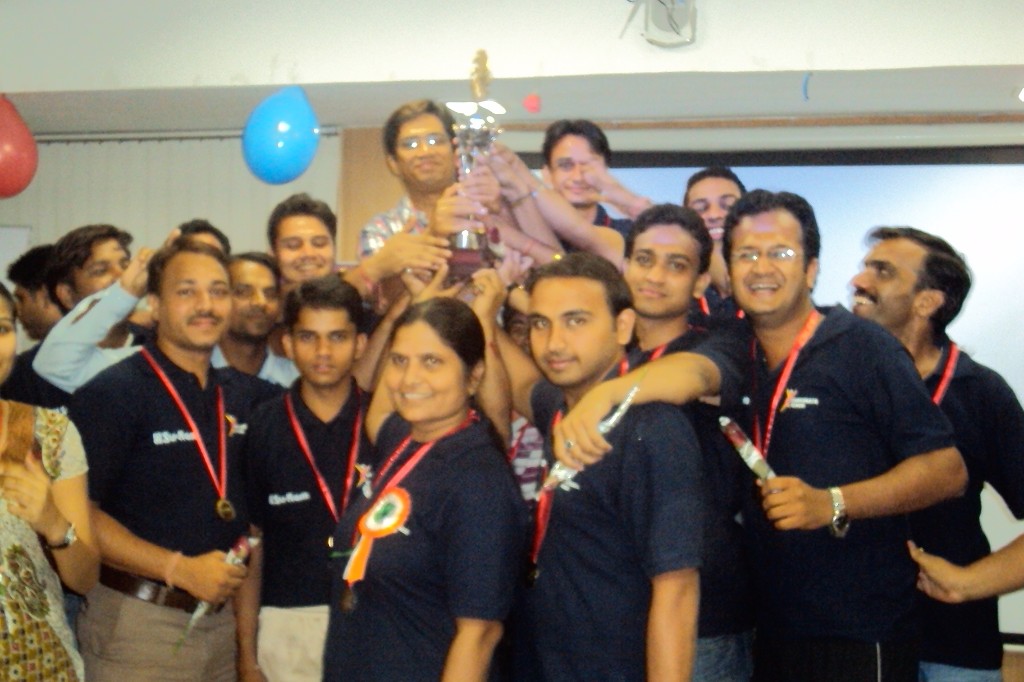 Su-Kam team in branded polos lifting a trophy indoors after Sports Week